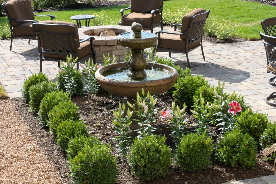 Tiered garden fountain in a planted patio bed with boxwood, lilies, and seating nearby